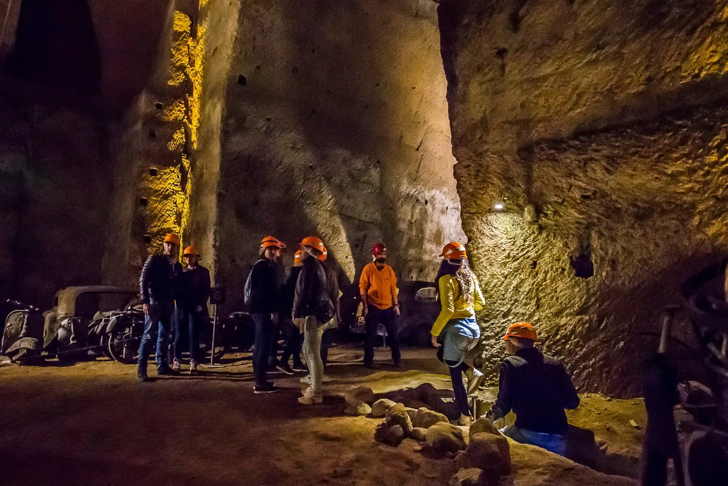 A group of visitors in orange hard hats standing in a lit cavern of the Bourbon Tunnel under Naples, cut through pale tuff with old vehicles parked against the far wall