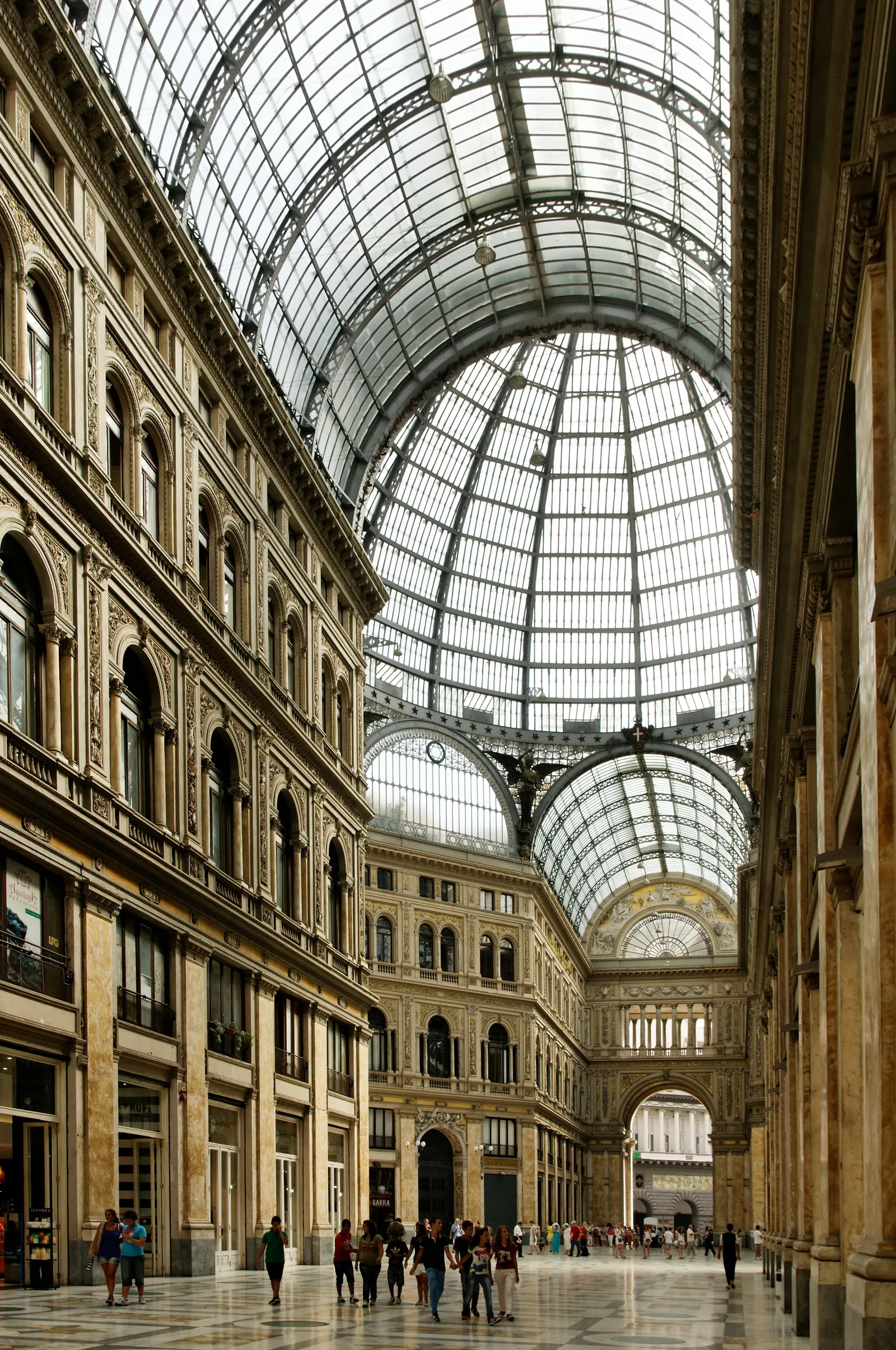Inside the Galleria Umberto I in Naples, a tall stone arcade under a barrel vault of iron and glass meeting a glazed dome at the crossing, with people walking the marble floor below