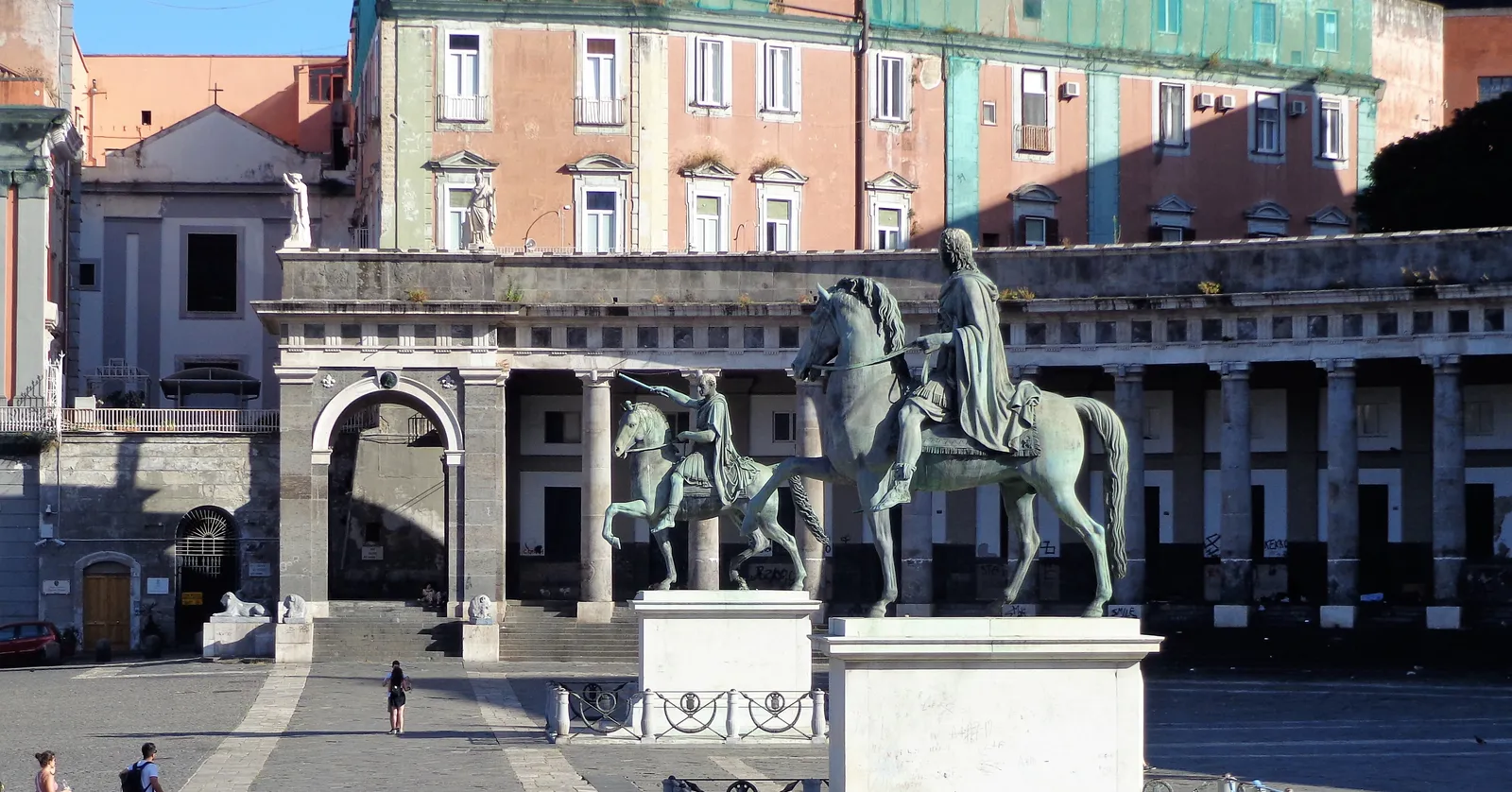Two bronze equestrian statues in the middle of Piazza del Plebiscito in Naples, with the curved colonnade of San Francesco di Paola running behind them