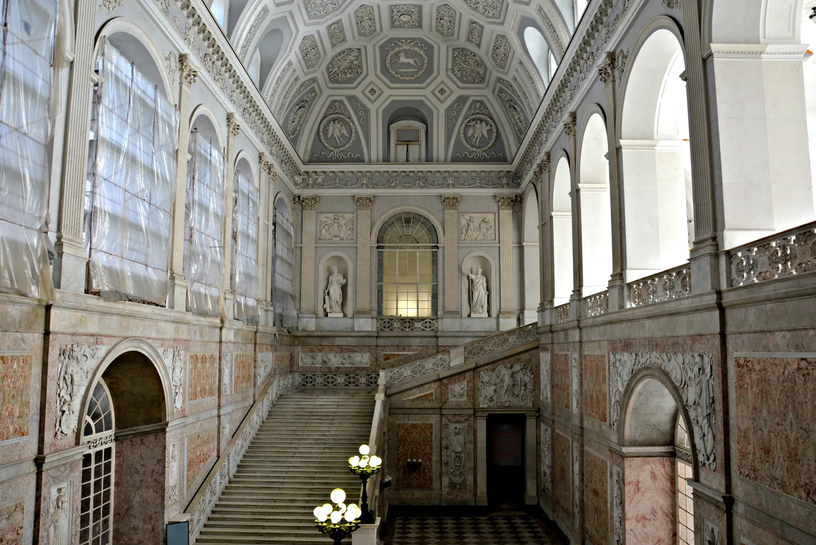 The grand staircase of the Royal Palace of Naples, a wide marble flight rising between coloured marble walls under a coffered vault with statues in niches