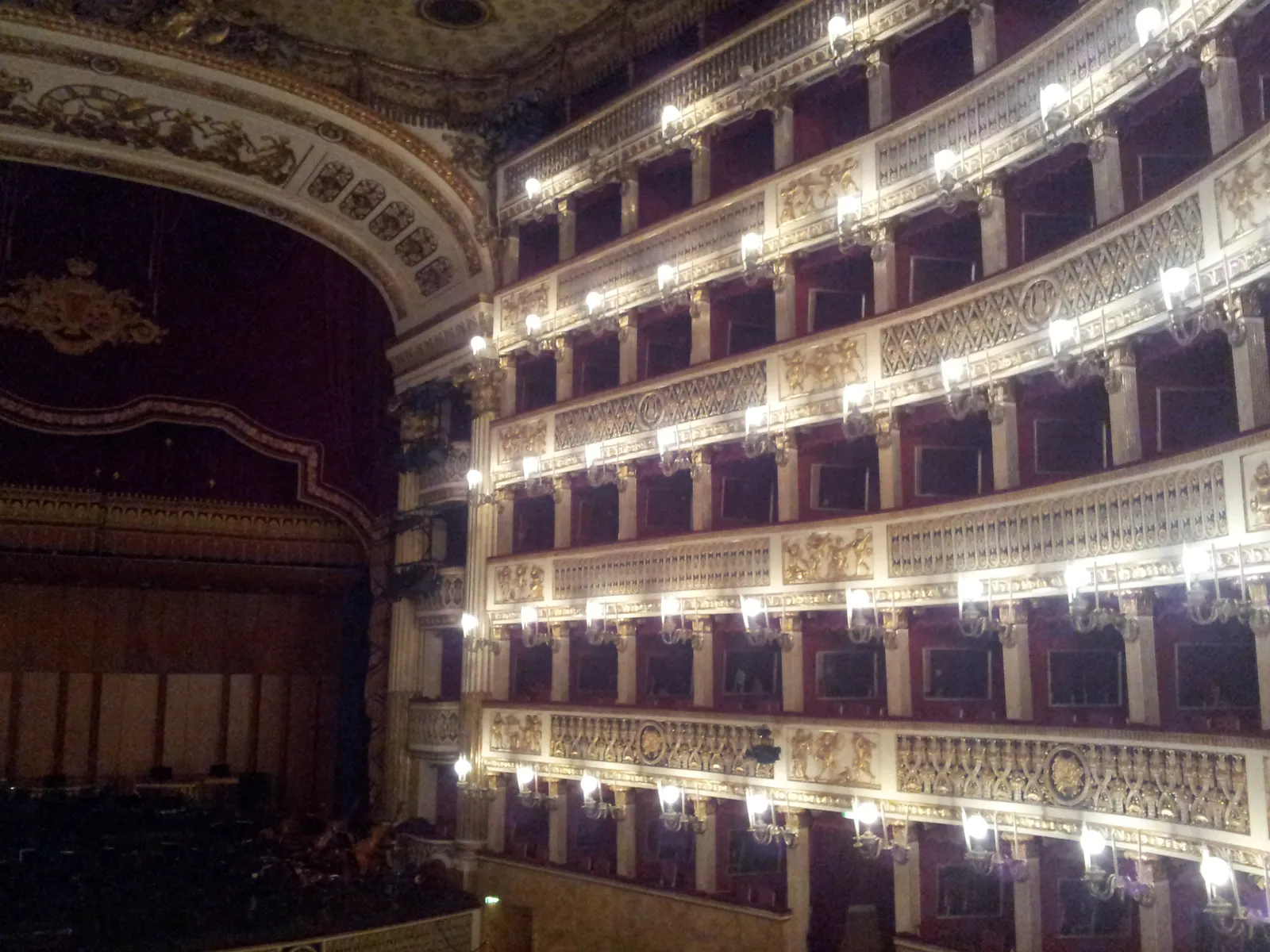 The auditorium of Teatro San Carlo in Naples, six curved tiers of gilded boxes lit by lamps, with the stage arch on the left