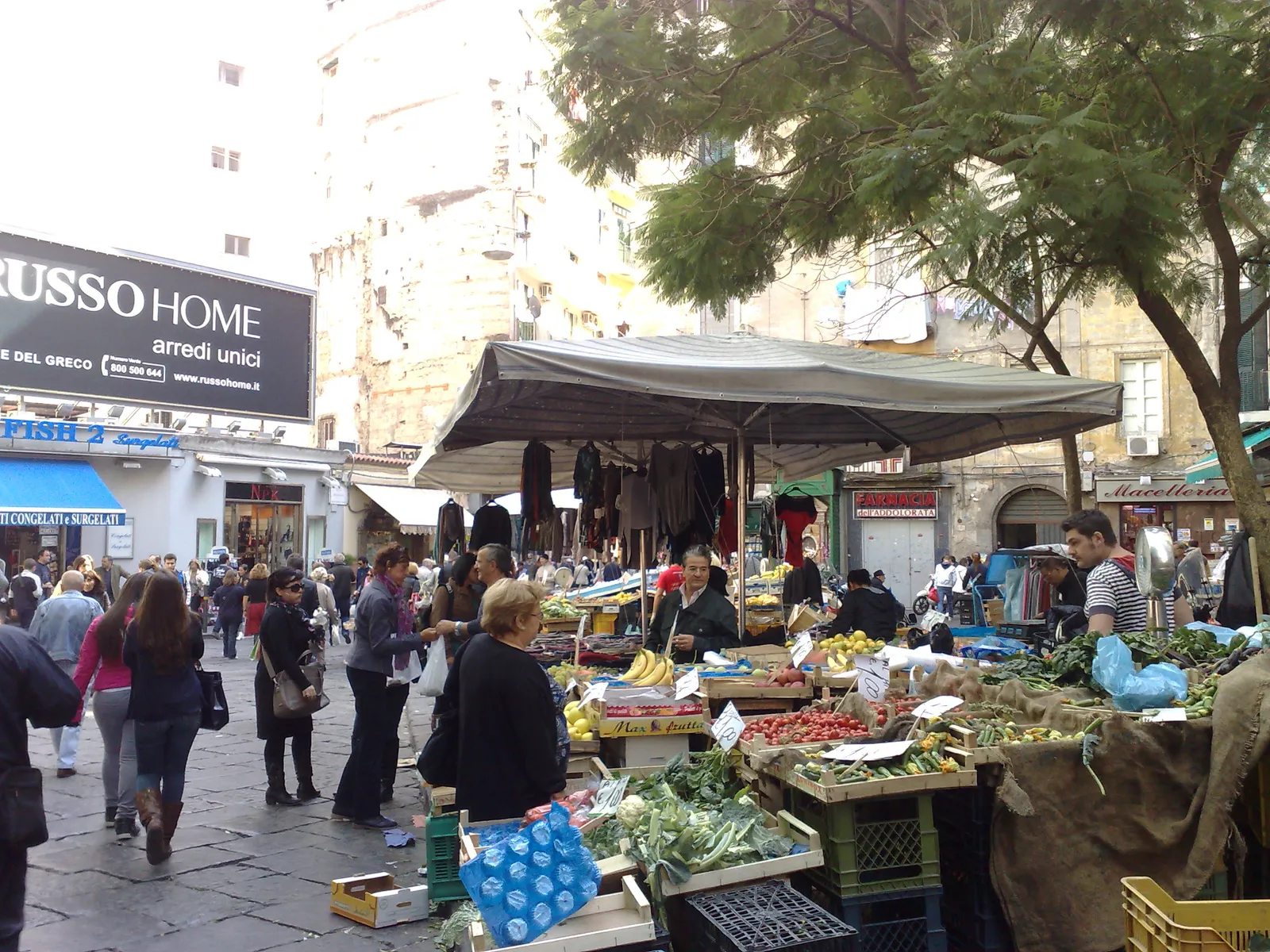 Stalls under canvas awnings at the Pignasecca market in Naples, piled with fruit and vegetables, with shoppers crowding the street between them