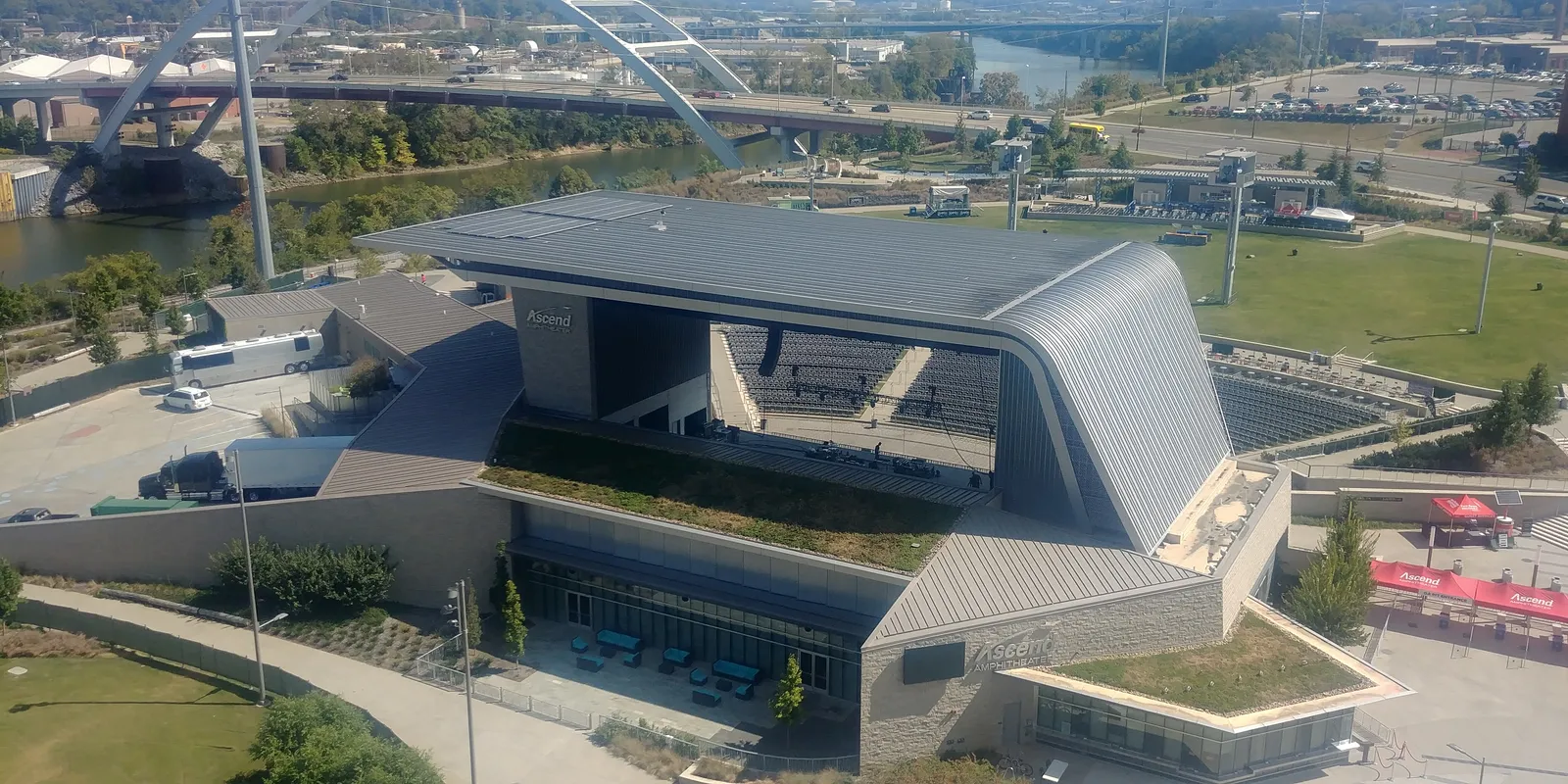 Ascend Amphitheater in Nashville from above, its angular metal canopy over a fan of fixed seats and a grass lawn, with the Cumberland River and bridges behind