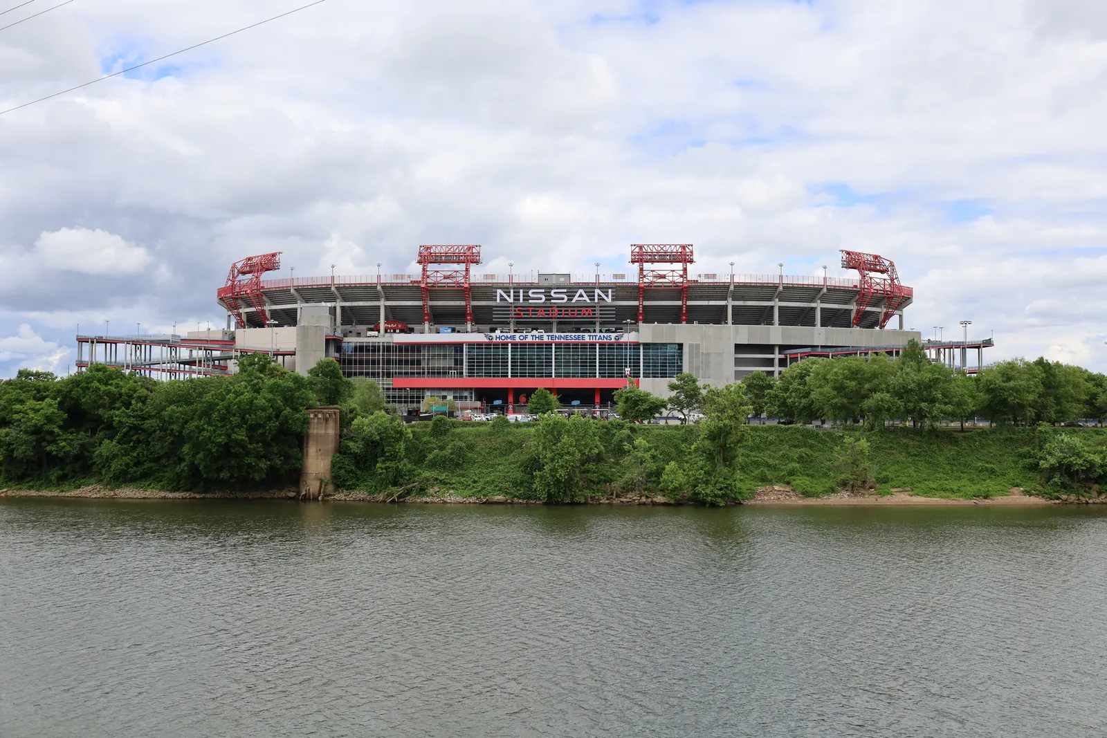The east bank of the Cumberland River in Nashville seen from the downtown side, with the football stadium and its red roof supports rising above the tree line