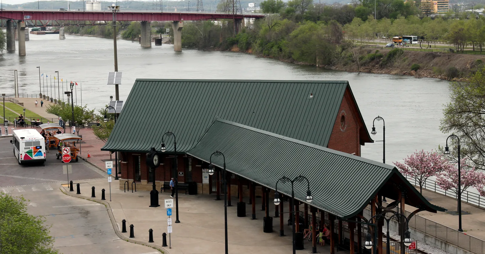 The green-roofed platform canopies of Riverfront Station in Nashville seen from above, with the Cumberland River behind them and a red steel road bridge crossing upstream