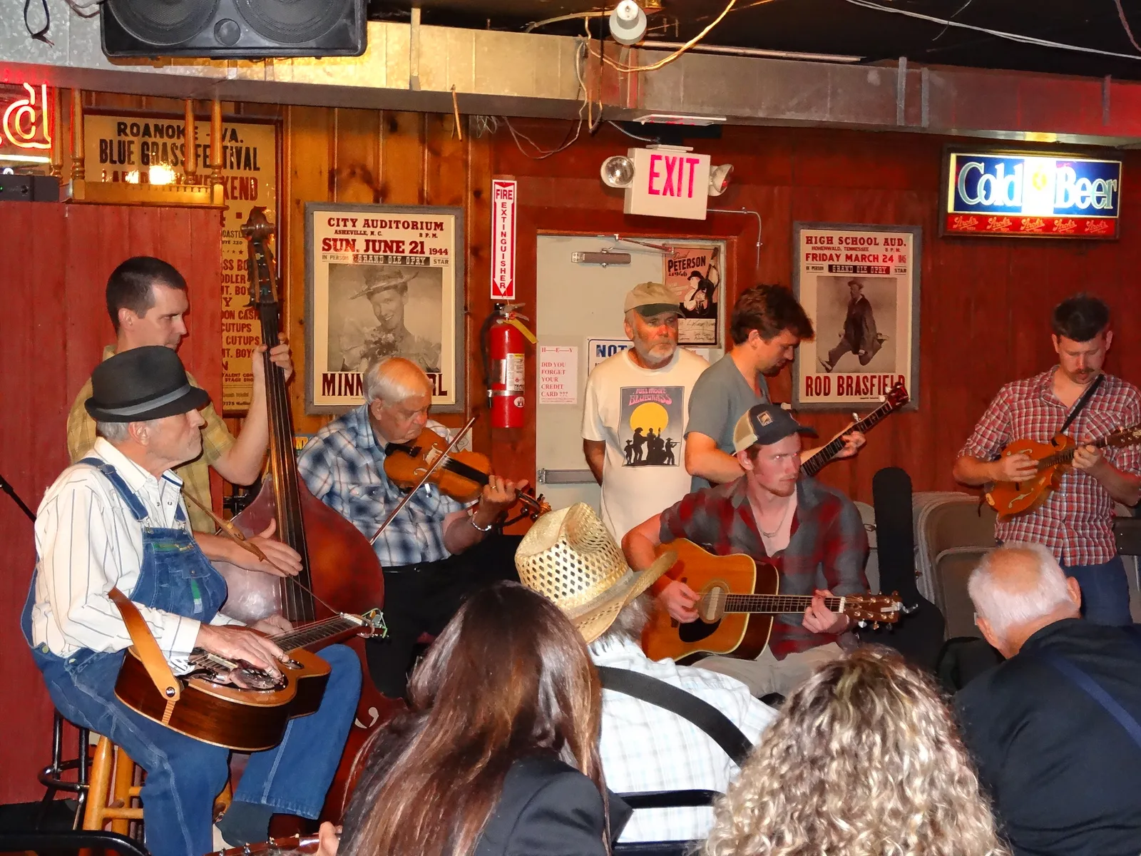 A bluegrass jam circle inside the Station Inn in Nashville, with players on guitar, fiddle, banjo and upright bass seated in a ring under old concert posters on wood-paneled walls