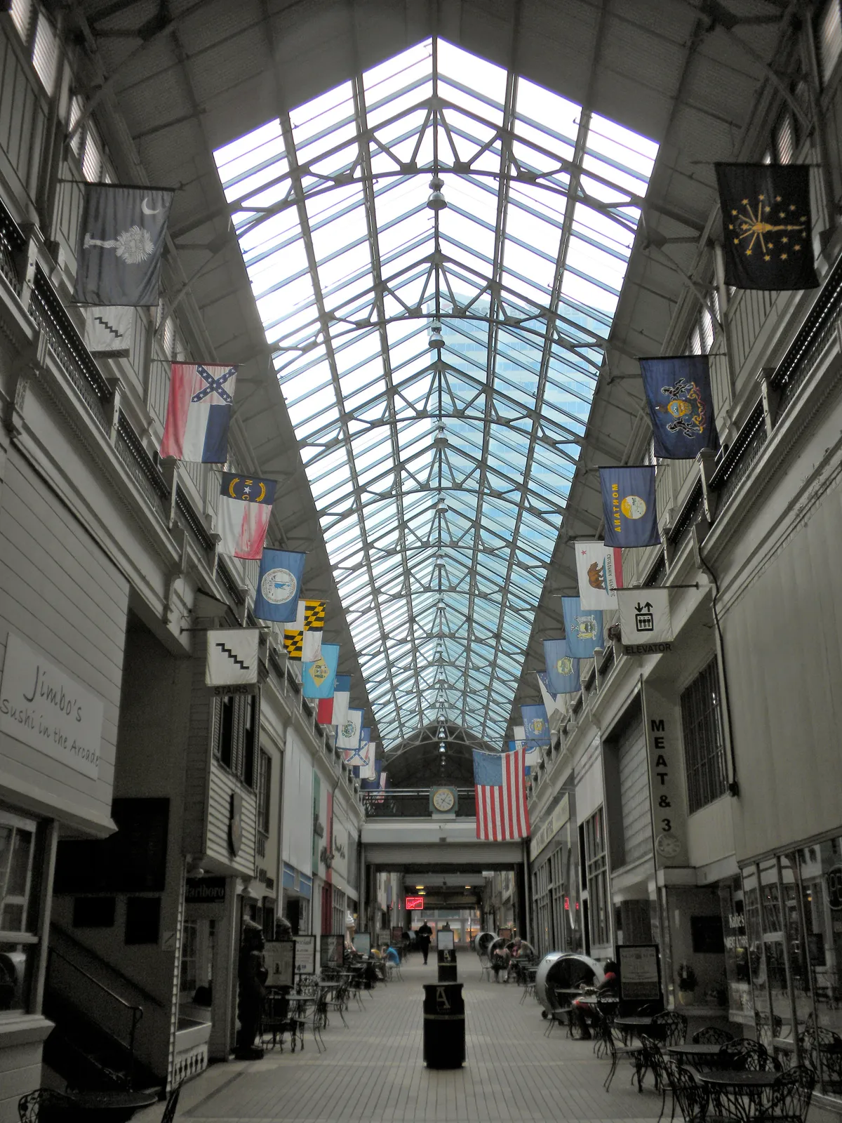 The interior of the Arcade in Nashville, a two-story gallery of shopfronts running under a gabled glass roof with flags hung along both upper balconies