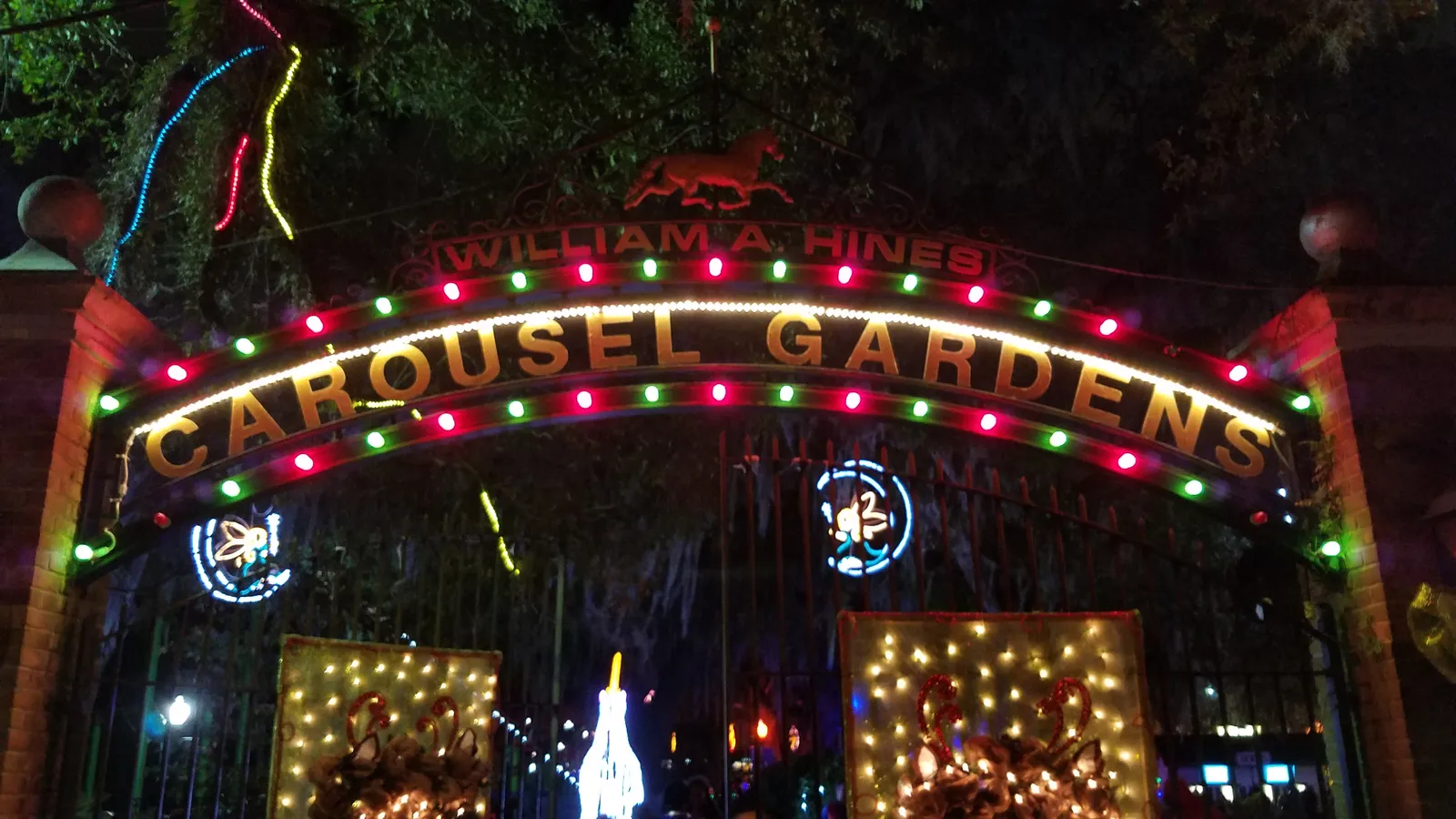 The entrance arch to Carousel Gardens in New Orleans City Park after dark, its lettering outlined in coloured bulbs with lit displays either side of the gate