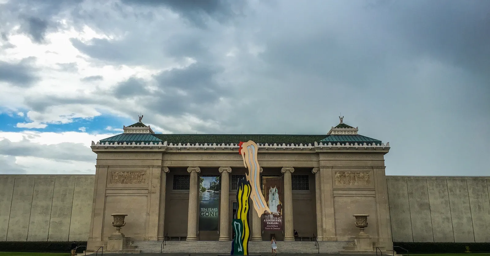 The front of the New Orleans Museum of Art, a pale stone Beaux-Arts building with an Ionic portico and low green-roofed wings, seen across its lawn under a heavy sky
