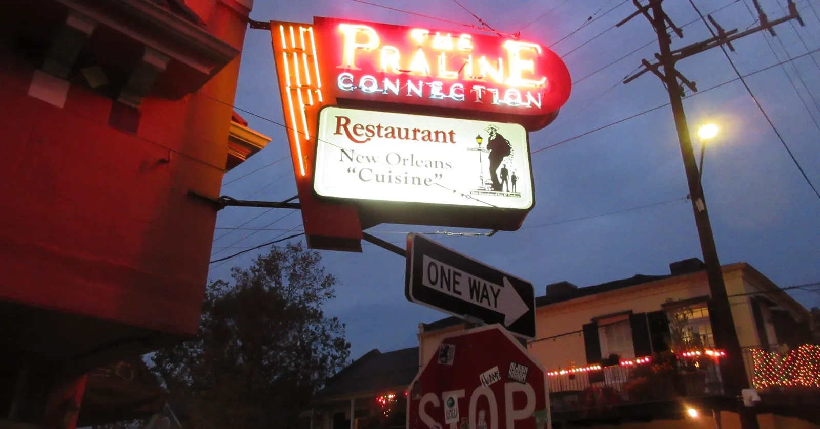 The corner of Frenchmen and Chartres Streets in New Orleans at dusk, a red and blue neon restaurant sign lit above the pavement with a one-way sign and low buildings behind