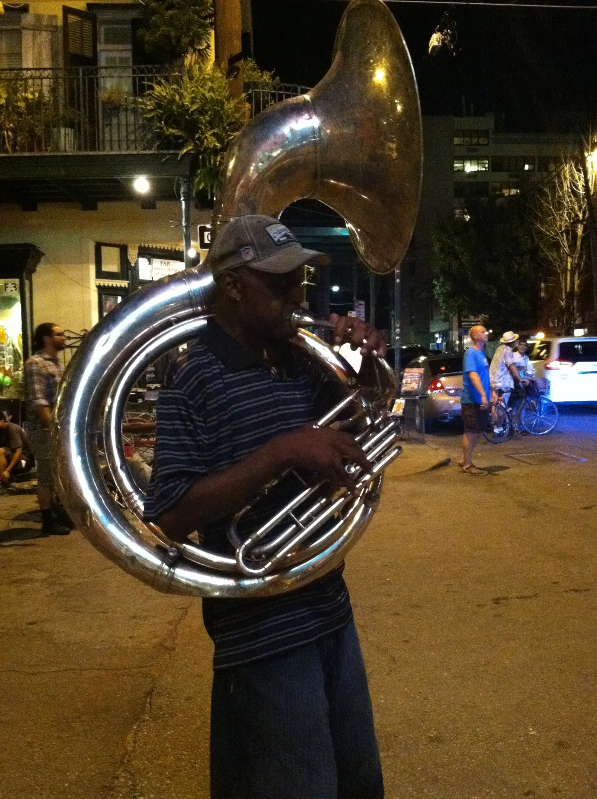 A street musician playing a silver sousaphone on the pavement of Frenchmen Street in New Orleans at night, with lit shopfronts and a small crowd behind him