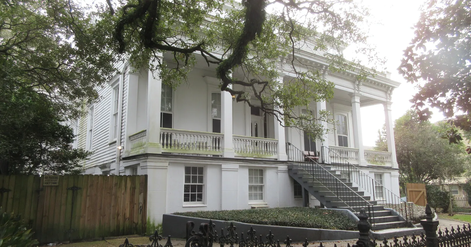 A white Greek Revival mansion on Prytania Street in the New Orleans Garden District, its two-storey gallery and double front staircase seen through the branches of a live oak, behind a black iron fence