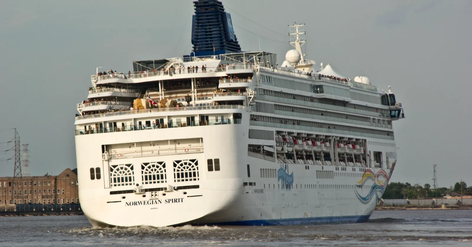 The stern of a large white cruise ship under way on the brown water of the Mississippi River at New Orleans, with low green riverbank behind it