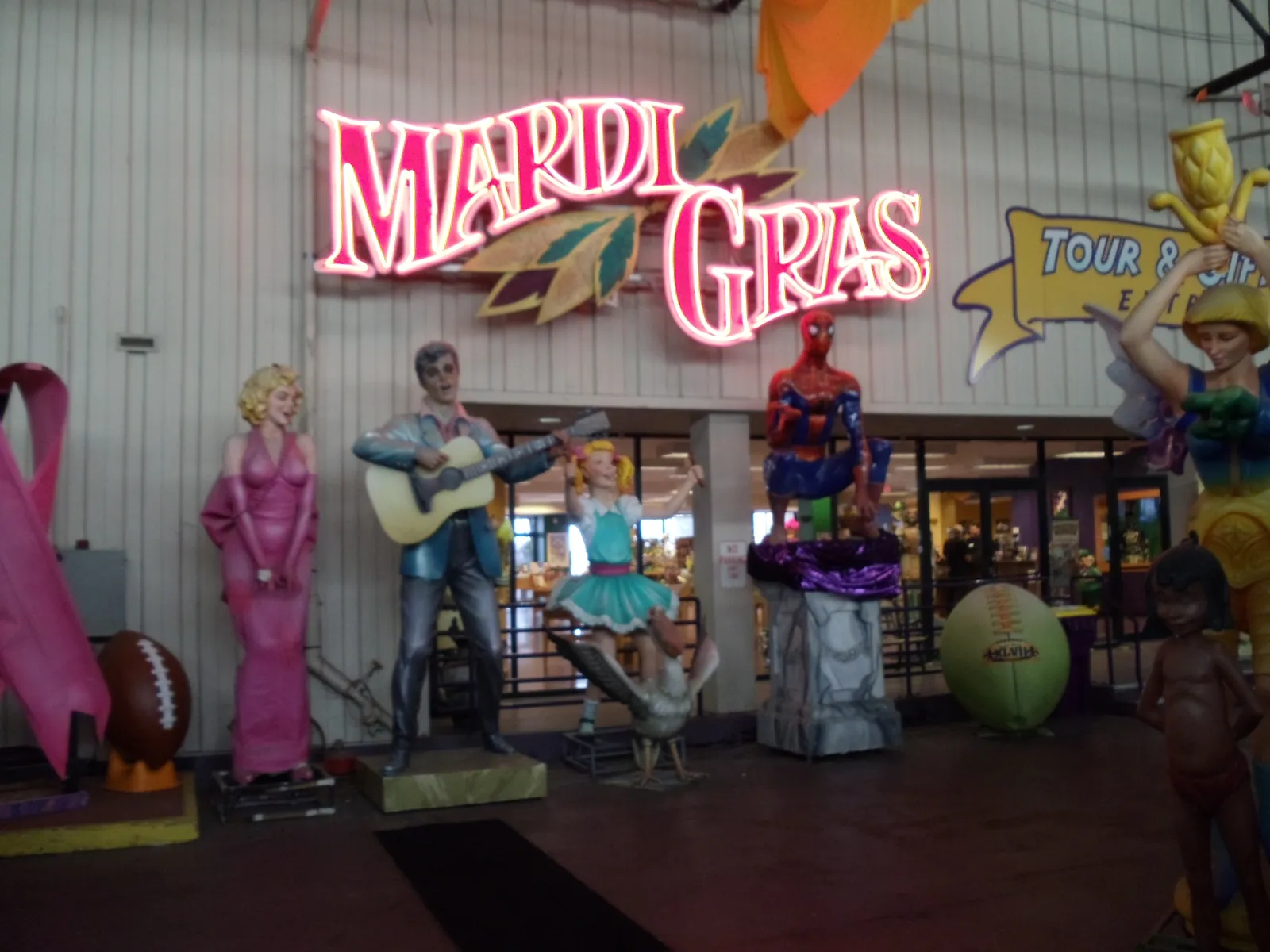 The entrance hall of Mardi Gras World in New Orleans, a red neon Mardi Gras sign on a corrugated warehouse wall above a row of painted parade figures