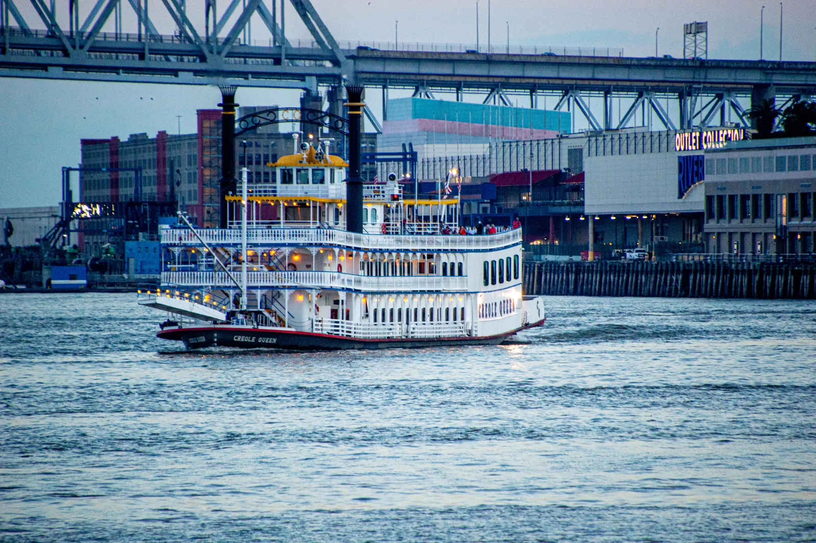 A white paddlewheel riverboat on the Mississippi at New Orleans passing the long wharf sheds of the Riverwalk, with the steel span of the Crescent City Connection bridge behind