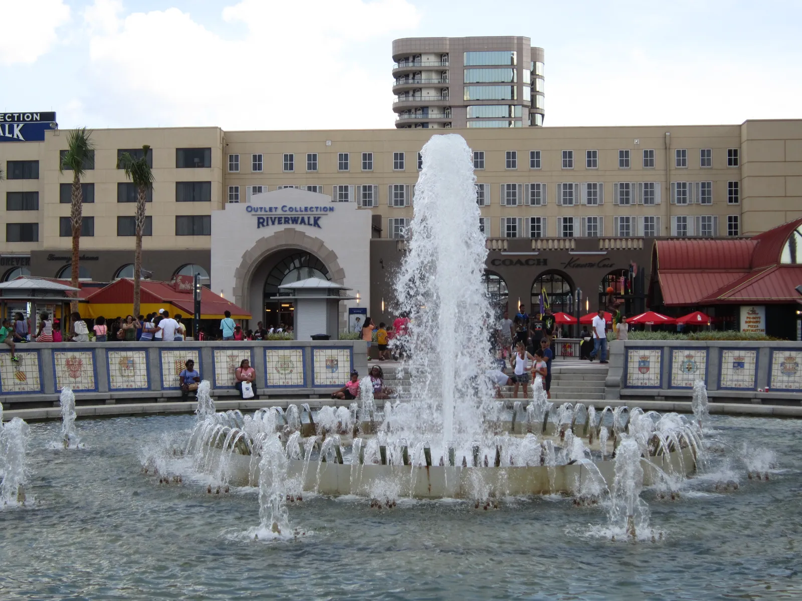 The circular fountain on Spanish Plaza in New Orleans throwing a tall central jet, its rim faced in painted heraldic tiles, with the Riverwalk shopping arcade behind