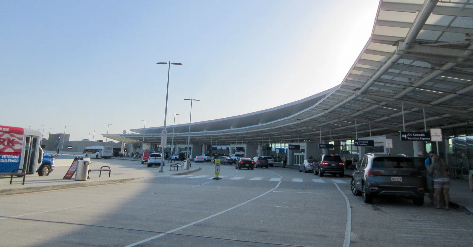 The kerbside of the terminal at Louis Armstrong New Orleans International Airport, a long white canopy curving away above the departures roadway with cars and passengers below it
