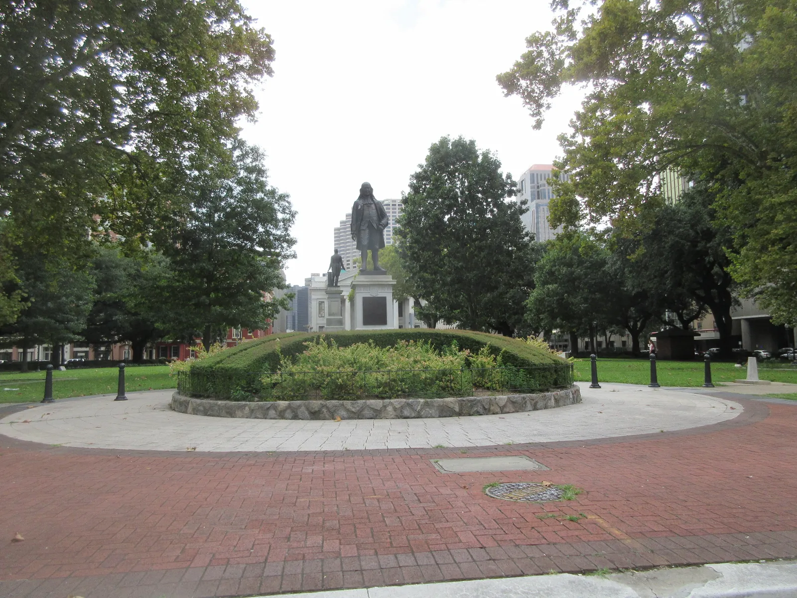 Lafayette Square in New Orleans, a bronze statue on a stone pedestal at the centre of a raised lawn, with brick paths and mature trees around it and office towers beyond