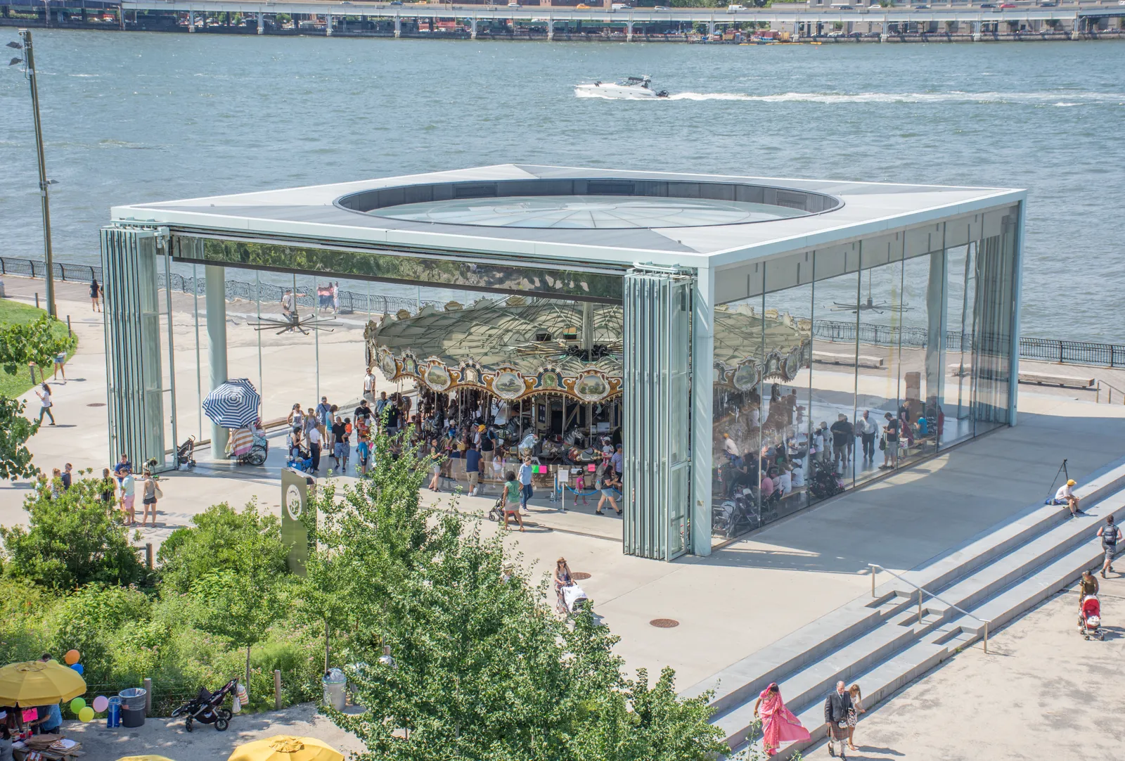 Jane's Carousel turning inside its glass pavilion on the Brooklyn Bridge Park waterfront, the East River and a passing boat behind it