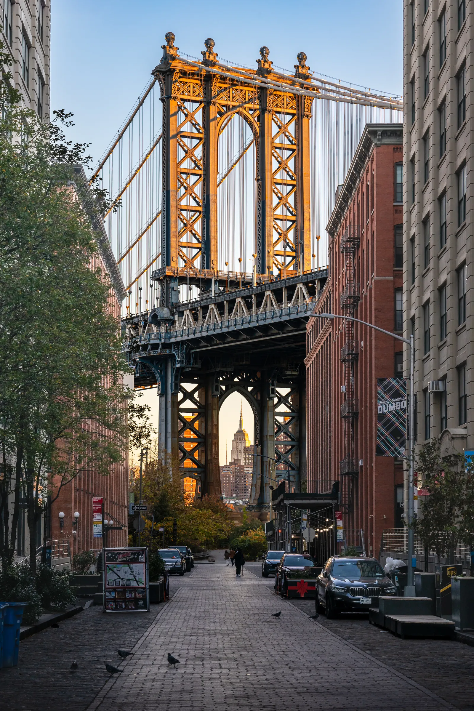 Washington Street in DUMBO paved in granite setts between brick warehouses, the Manhattan Bridge tower spanning the gap above with the Empire State Building framed underneath it