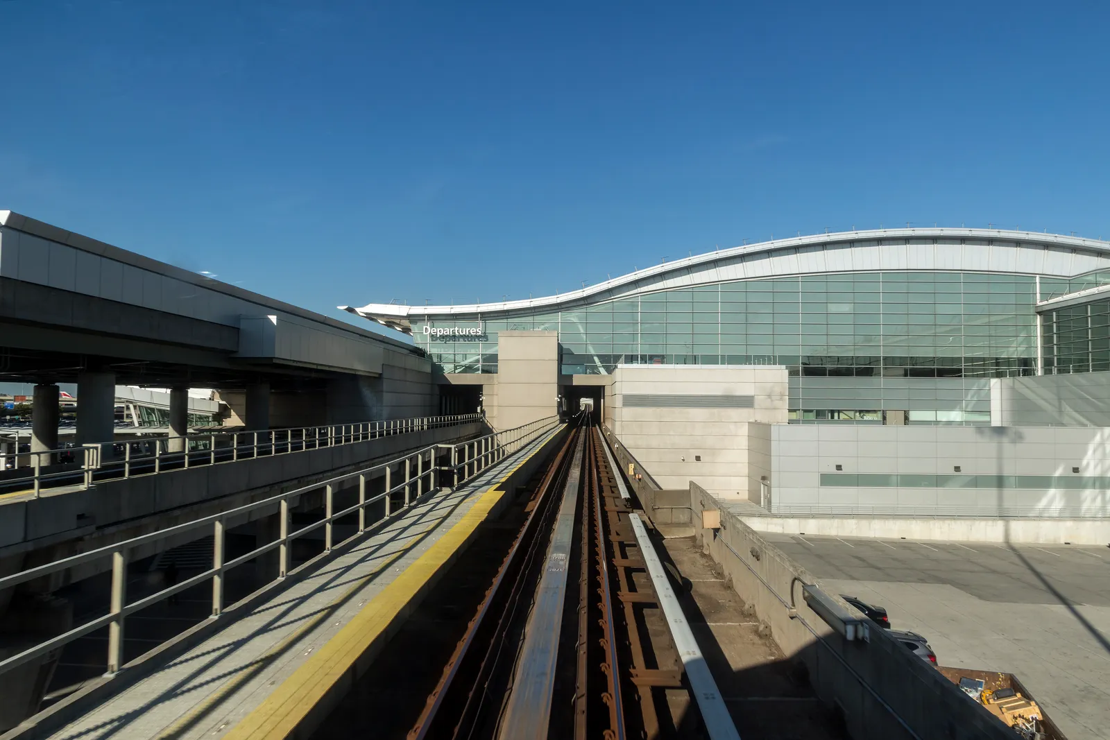 The elevated AirTrain guideway at JFK Airport running toward the glass frontage of Terminal 4, with a platform and Departures signage ahead