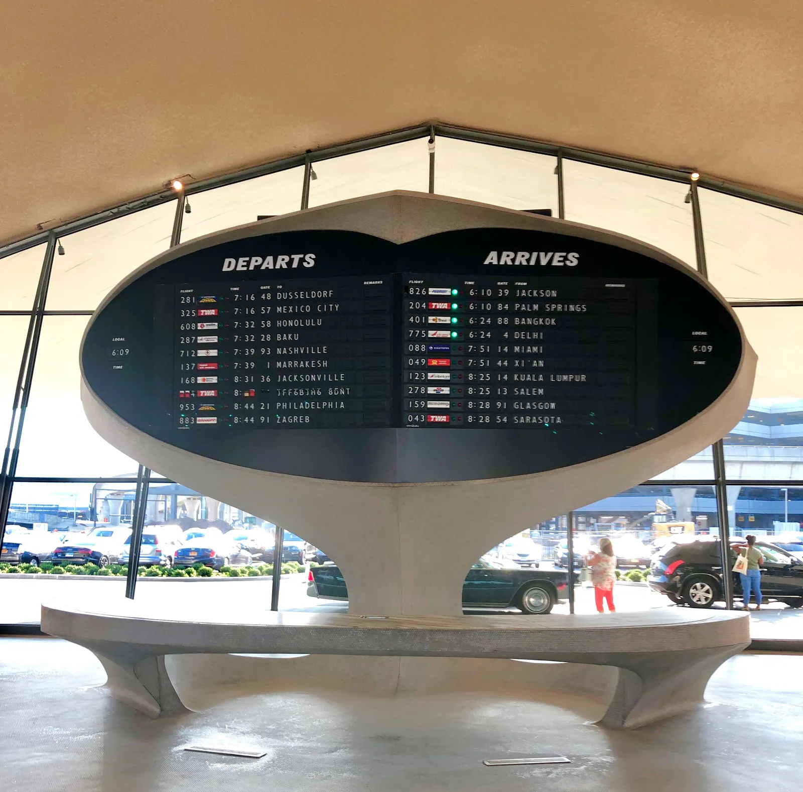 A black split-flap departures and arrivals board mounted on a white concrete pedestal inside the TWA Flight Center, with a curved bench running around its base