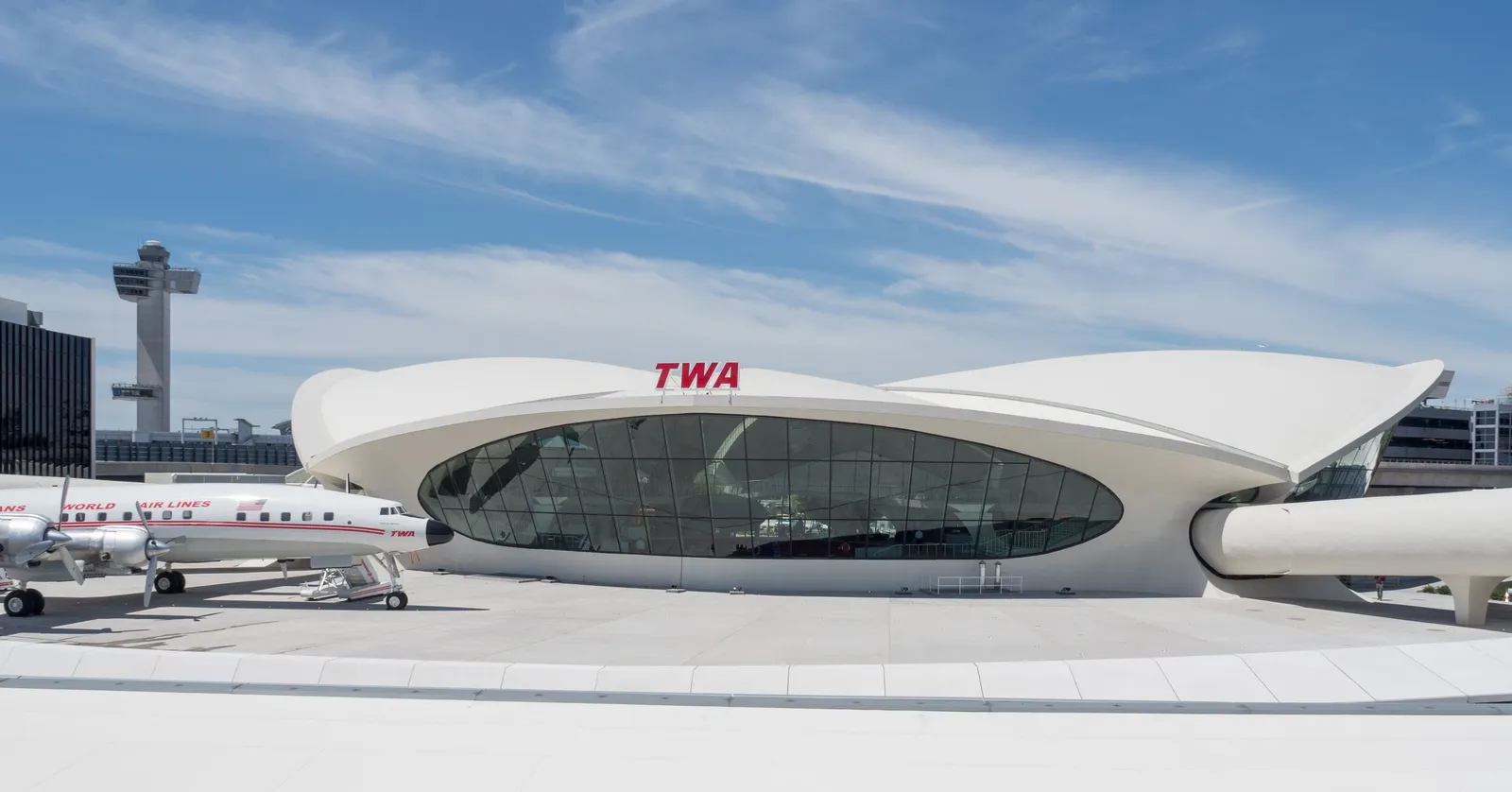 The white concrete TWA Flight Center at JFK Airport under a blue sky, its winged roof spread over a curved glass front with TWA lettering, a propeller airliner parked at the left