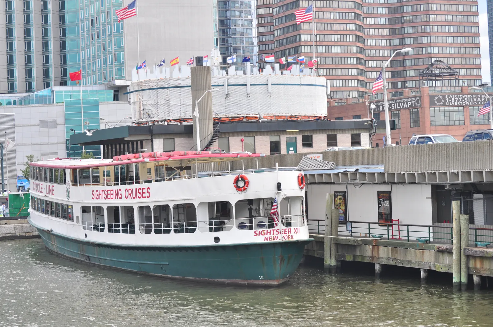 A Circle Line sightseeing boat with a white upper deck and green hull moored at its dock in Manhattan, with flags flying on the terminal building behind