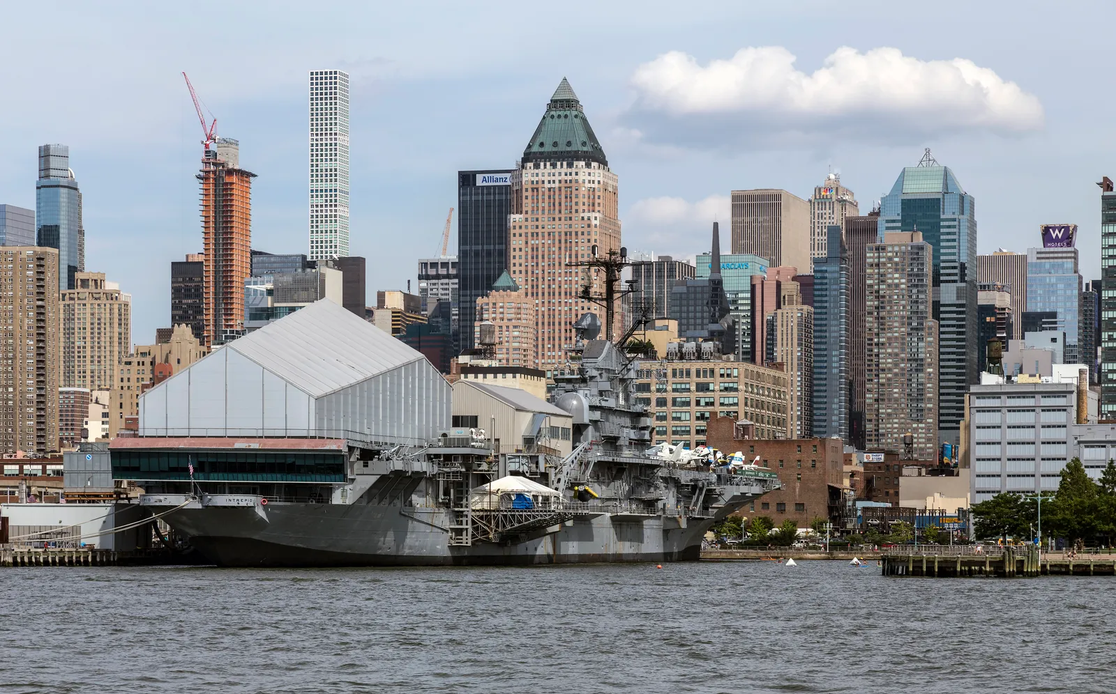 The grey aircraft carrier Intrepid moored at Pier 86 on the Hudson River in Manhattan, with the white Space Shuttle Pavilion on her flight deck and Midtown towers rising behind