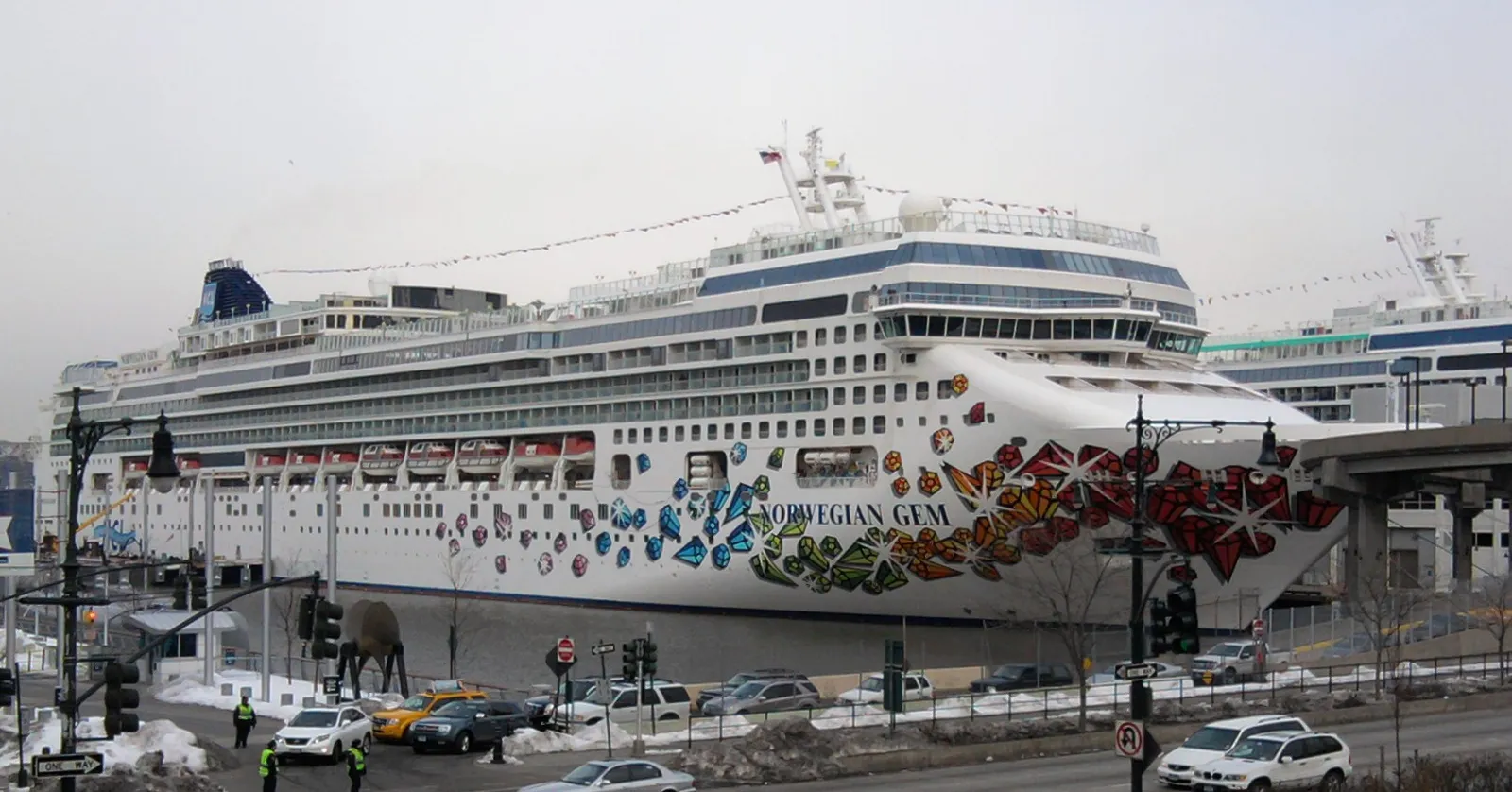 A white cruise ship painted with bright jewel shapes on her bow, tied up at Pier 88 of the Manhattan Cruise Terminal beside Twelfth Avenue in New York, with taxis on the street below