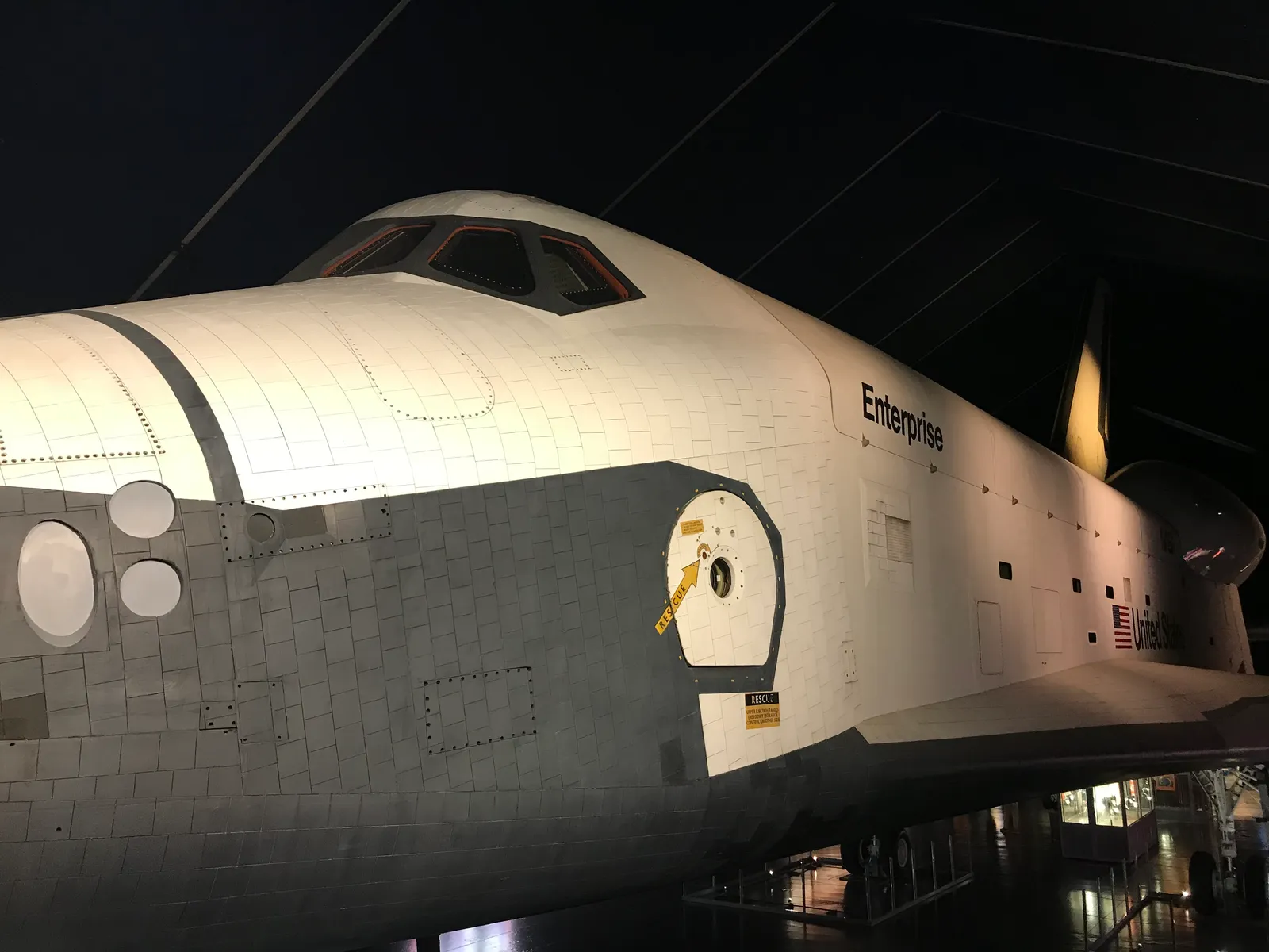 The nose and cockpit windows of the Space Shuttle Enterprise, its name painted on the white fuselage, lit up inside the darkened Space Shuttle Pavilion at the Intrepid Museum in New York