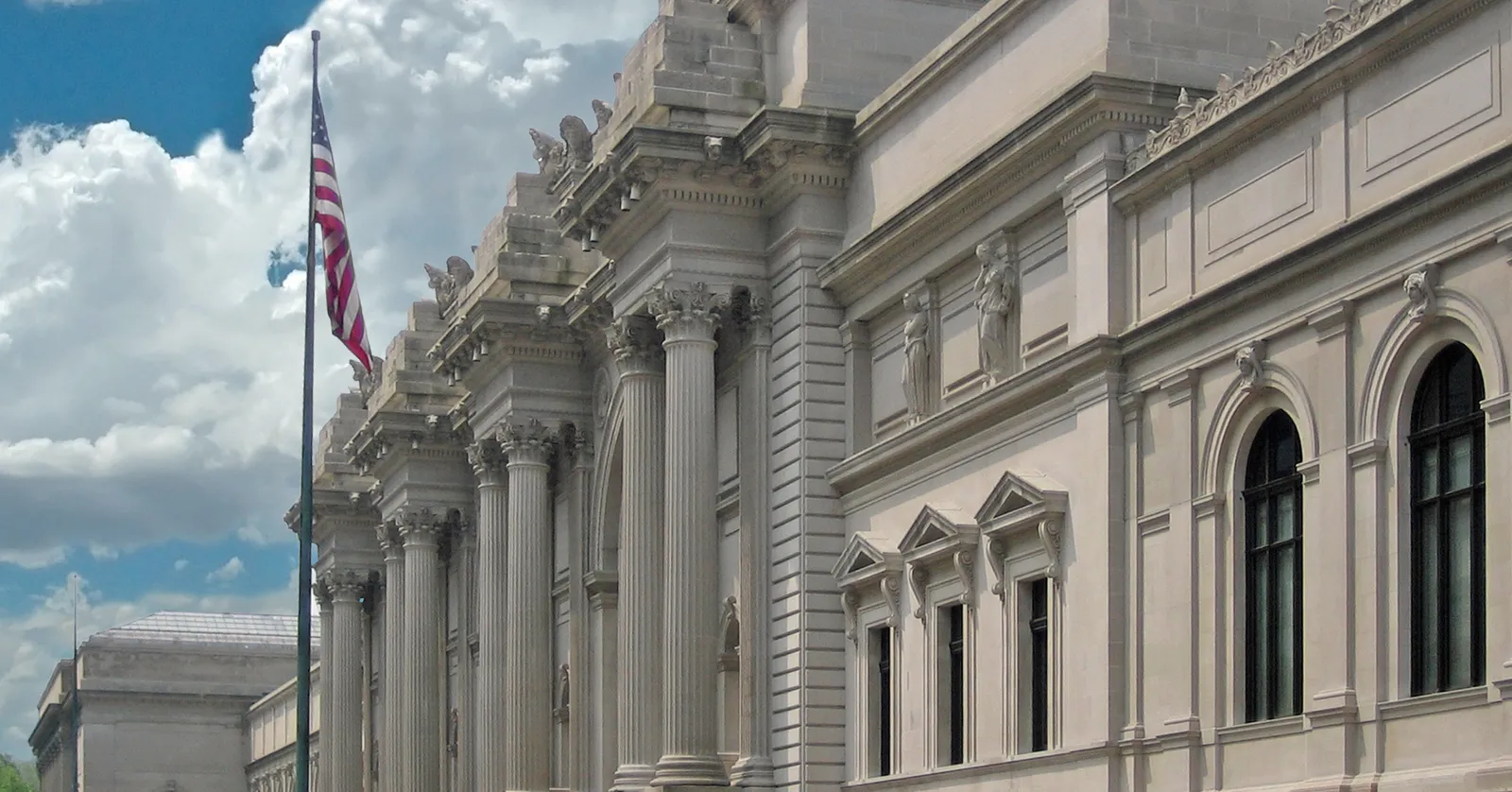 The Fifth Avenue front of the Metropolitan Museum of Art, a long limestone facade of paired columns and arched windows with flags flying from poles along the pavement