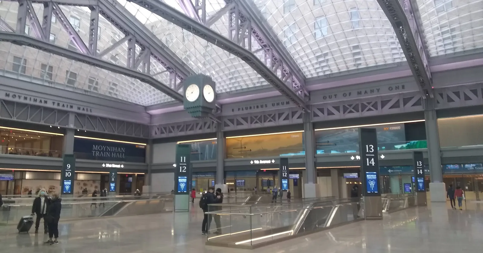 The main floor of Moynihan Train Hall in New York, riveted steel trusses carrying four glass vaults over the concourse, a four-faced clock hanging in the middle and platform number columns below