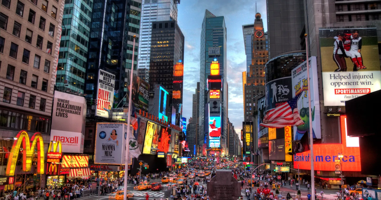 Times Square in New York looking south at dusk, illuminated advertising screens stacked up both sides of the street and crowds filling the pedestrian plazas below