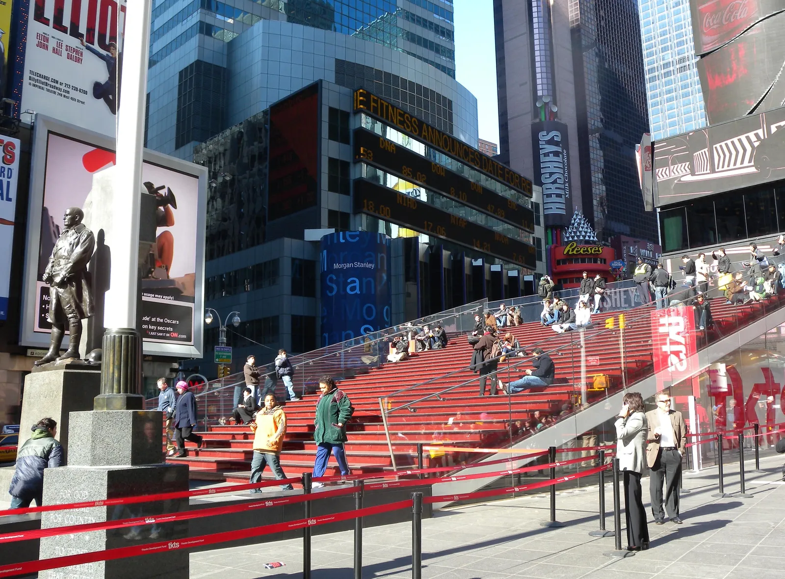 The bank of red glass steps above the TKTS ticket booth in Father Duffy Square, people sitting on them in the sun with the Father Duffy statue in the foreground