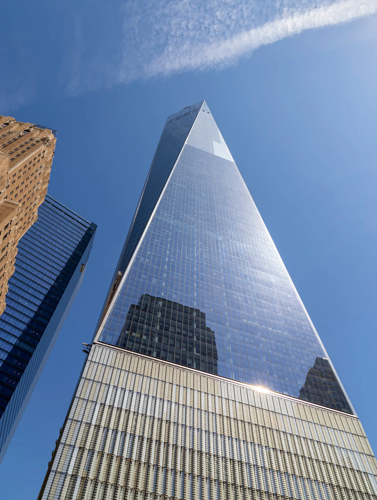 One World Trade Center seen from the street below, its glass sides tapering into eight triangles toward the top with the mast above them against a clear sky