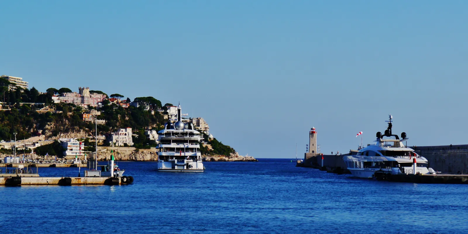 The entrance to Port Lympia in Nice, with a small lighthouse, moored yachts, and the wooded slope of Mont Boron behind