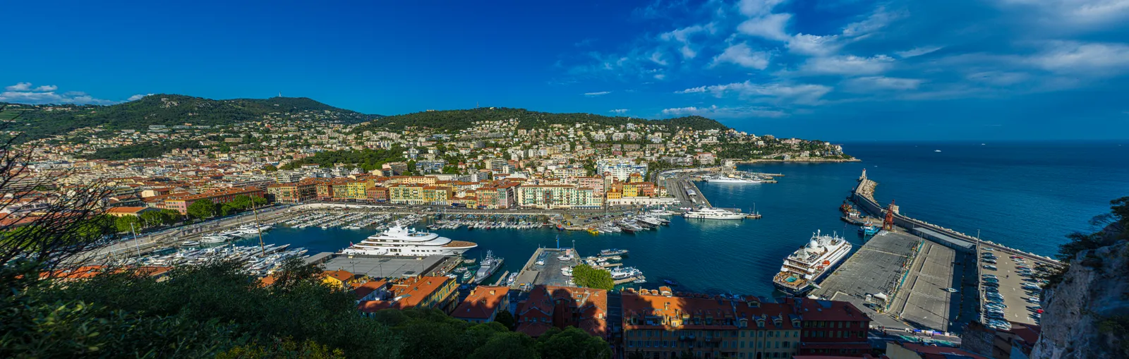 An elevated panoramic view of Port Lympia in Nice, showing the harbor's colorful buildings, moored yachts, and the surrounding hillside