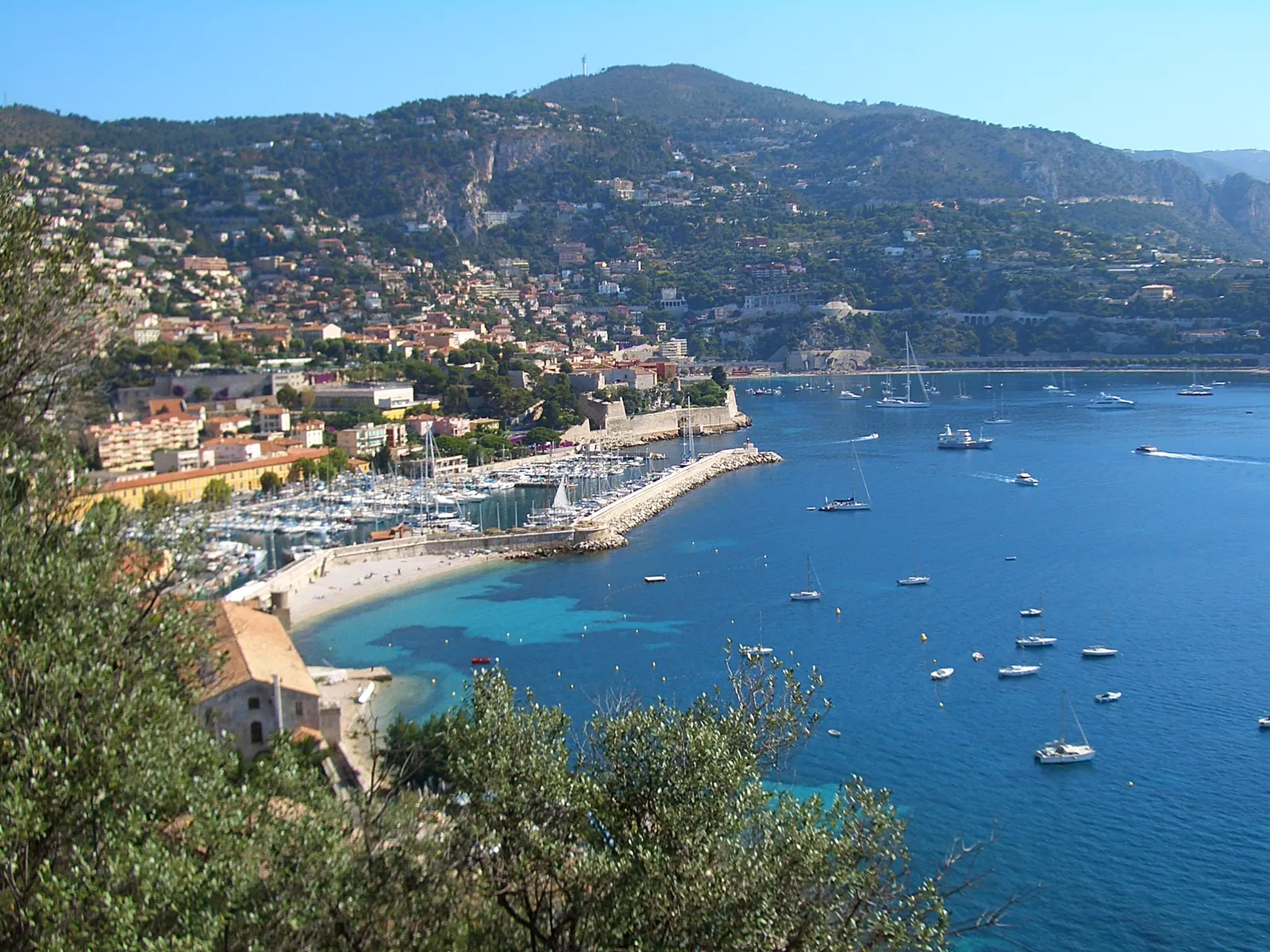 An elevated view of Villefranche-sur-Mer's bay, showing the citadel, marina, sandy beach, and turquoise water framed by hills