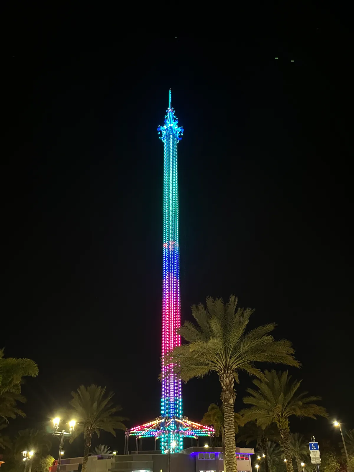The Orlando StarFlyer tower lit in green, blue and pink after dark, its swing gondola at the base ringed by palm trees on International Drive