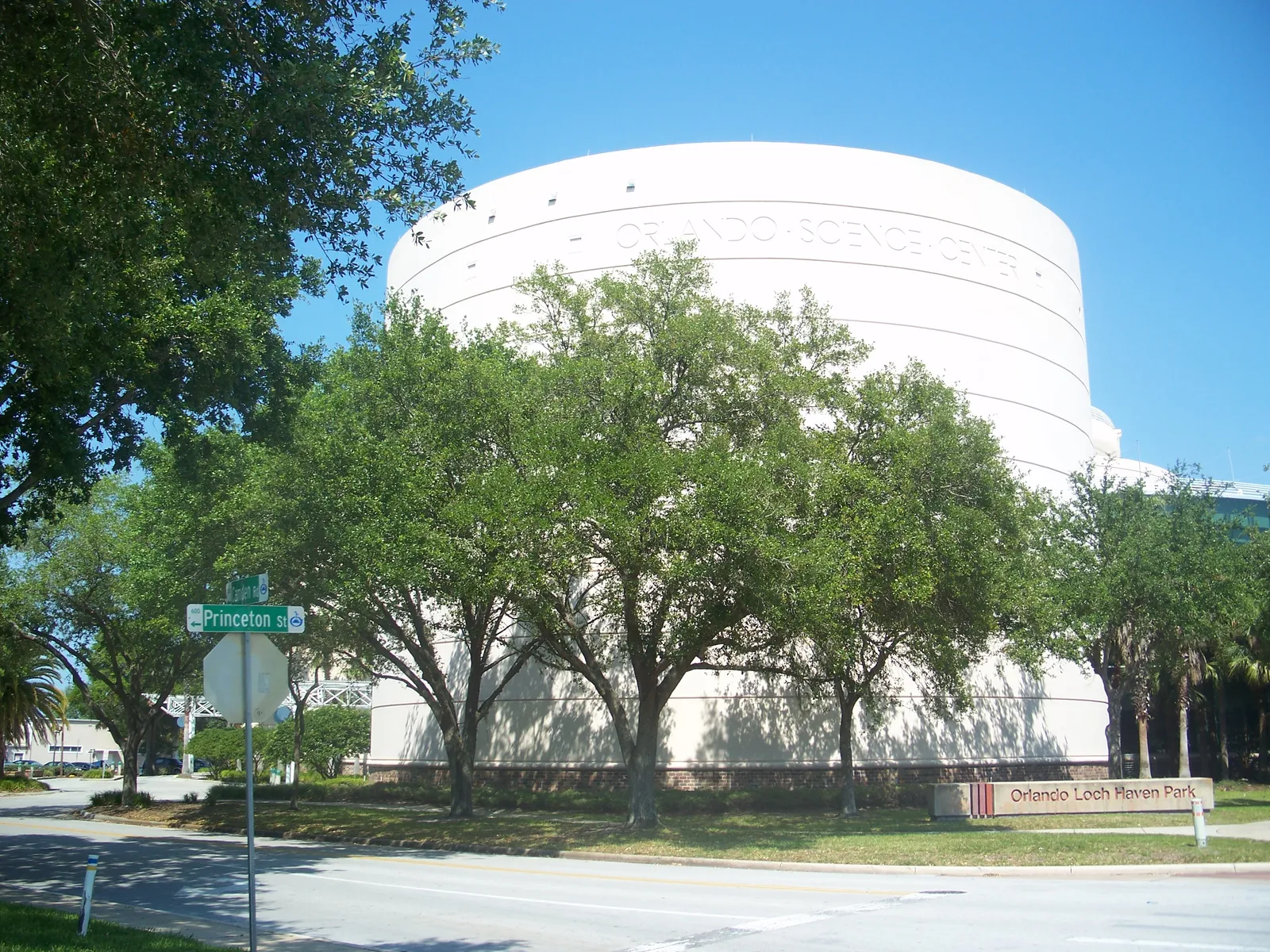 The white cylindrical drum of the Orlando Science Center rising behind oak trees at the corner of Princeton Street in Loch Haven Park