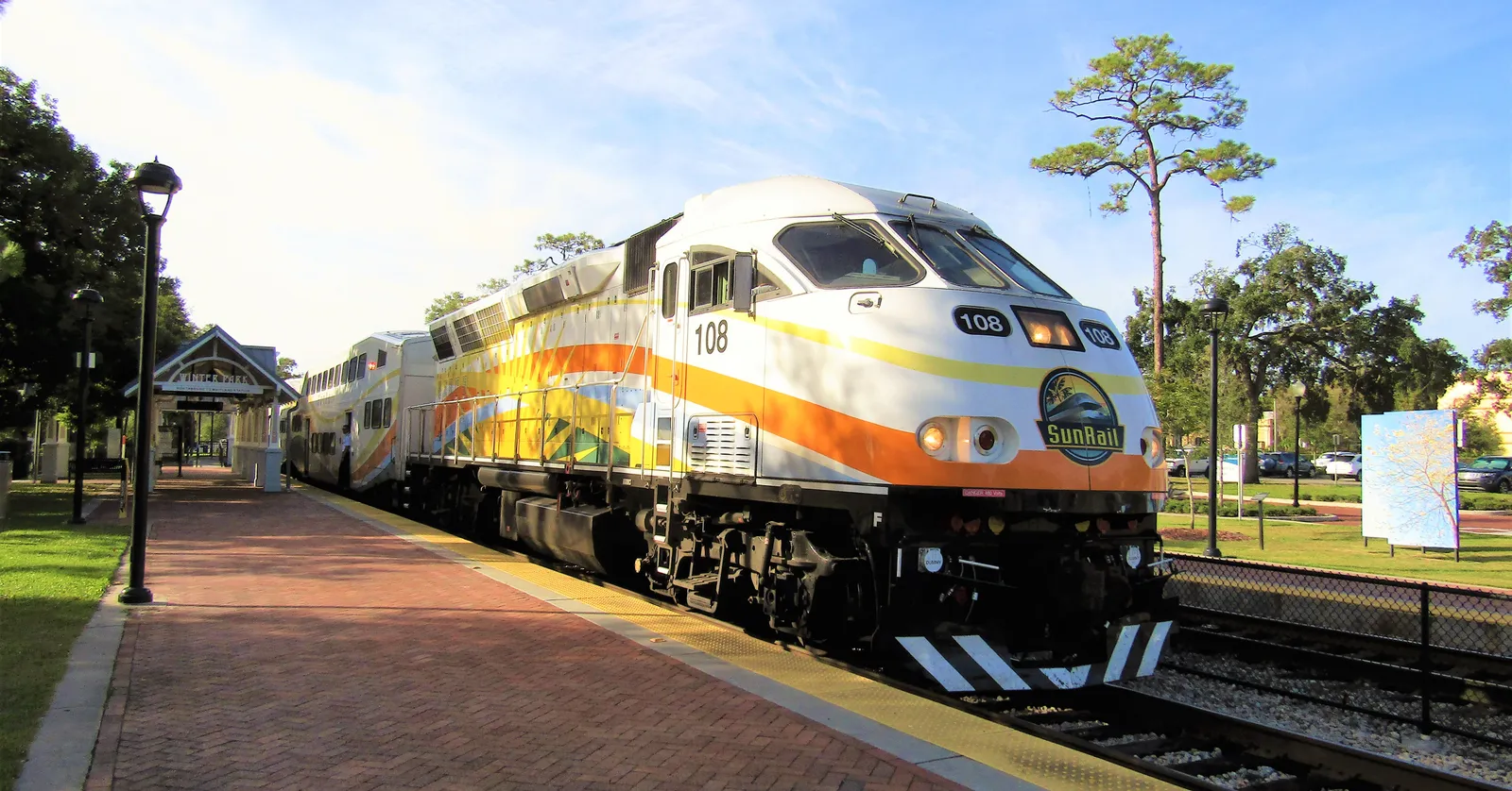 A SunRail train standing at Winter Park station in Florida, with the white gabled roof of the Craftsman-style depot and its brick platform alongside