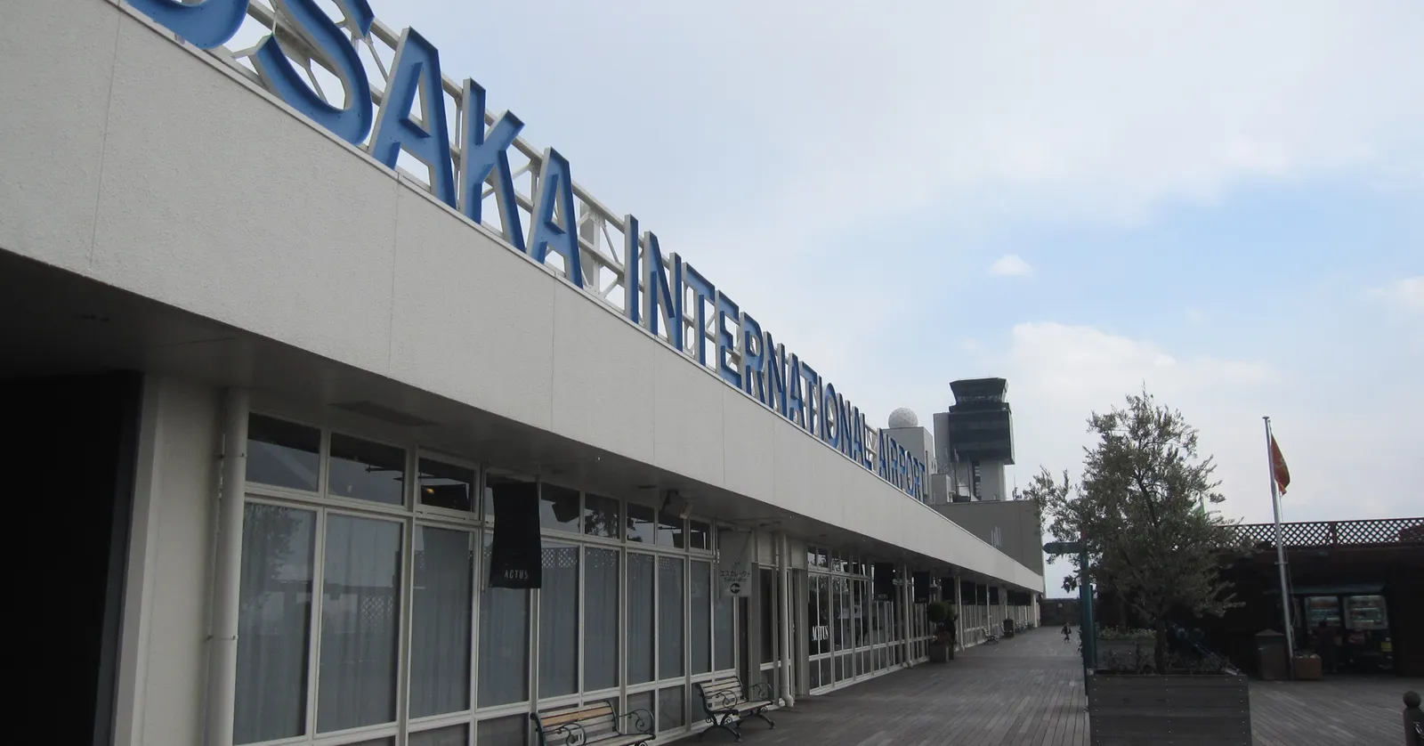 The kerbside frontage of Osaka International Airport at Itami, a long low terminal with OSAKA INTERNATIONAL AIRPORT spelled out in tall blue letters along the roof edge and the control tower standing behind it