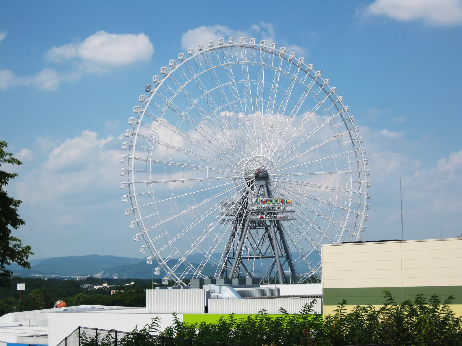 The Redhorse Osaka Wheel at Expocity, a white steel Ferris wheel with a full ring of enclosed gondolas standing against a blue sky above low buildings and trees