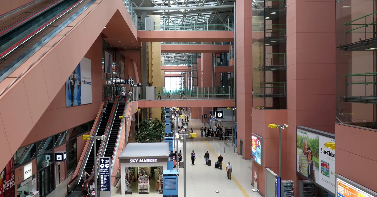 The long glass-roofed hall of Terminal 1 at Kansai International Airport near Osaka, with escalators, walkways and travelers with luggage below