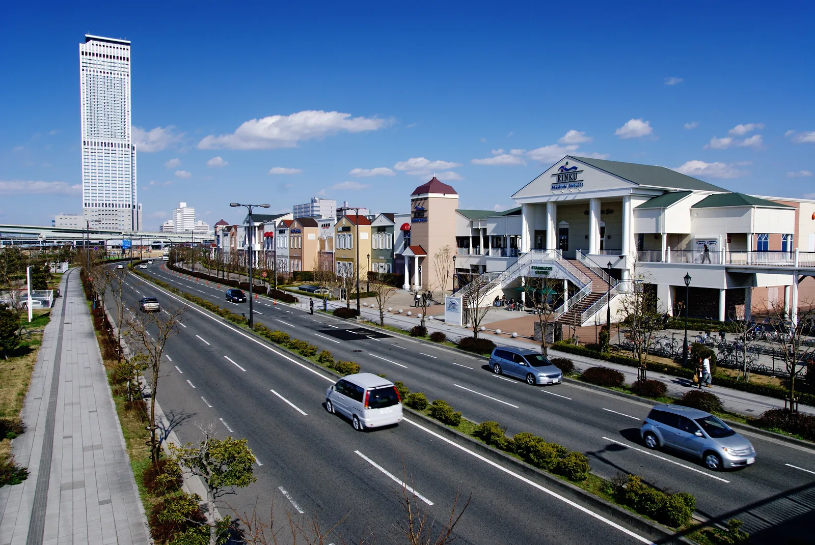The low colonial-style buildings of Rinku Premium Outlets in Rinku Town near Kansai Airport, with the tall Rinku Gate Tower Building behind under a blue sky
