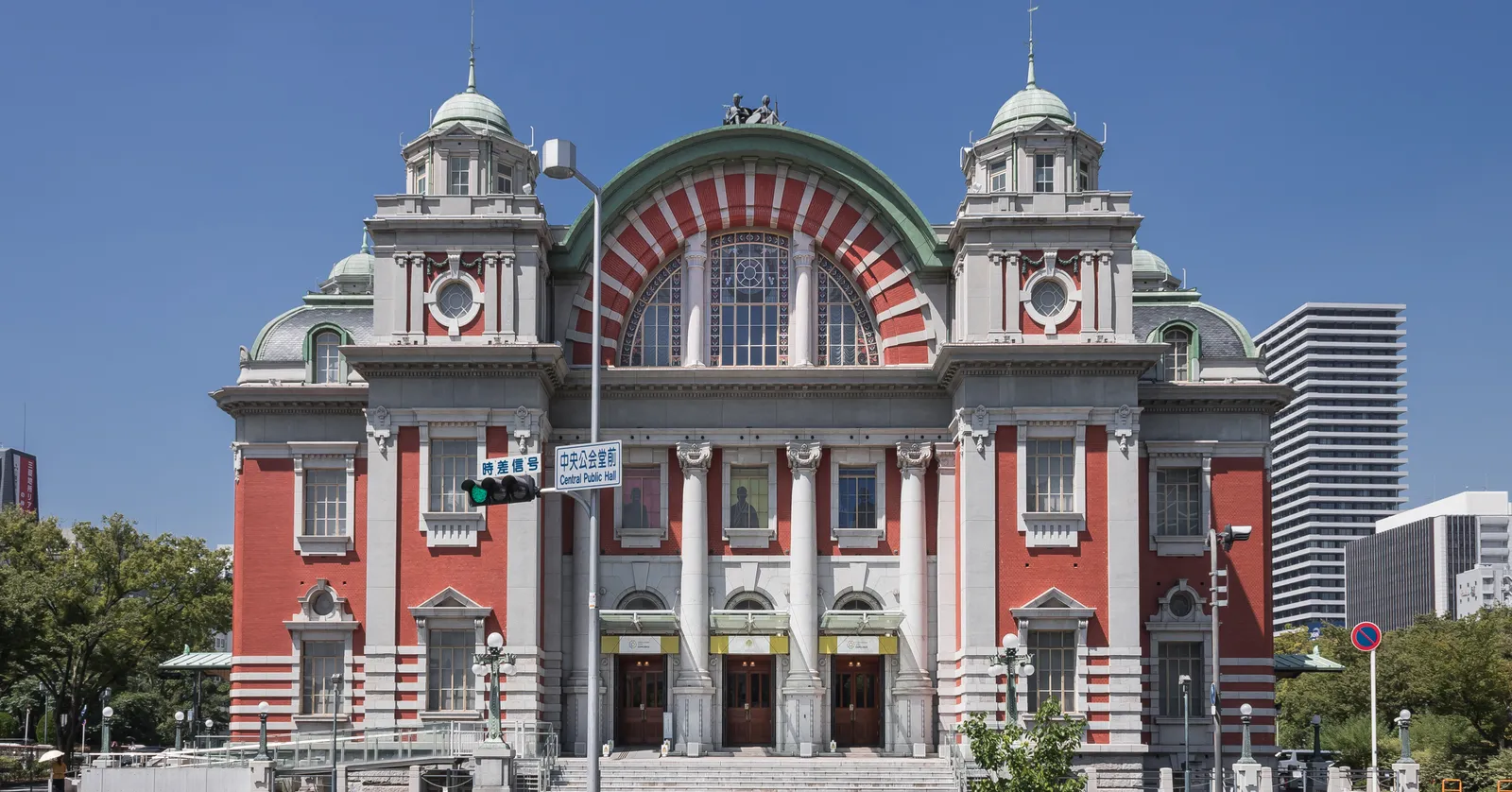 The front of Osaka City Central Public Hall, a symmetrical red-brick and pale stone building with a wide arched window under a green copper barrel roof and a domed turret at each corner