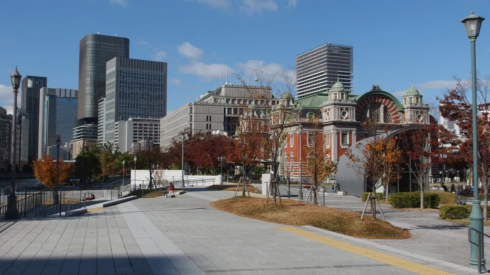 Nakanoshima Park in Osaka in autumn, a wide paved terrace with young trees in red leaf, the green-domed roof of the Central Public Hall to the right and office towers along the skyline behind