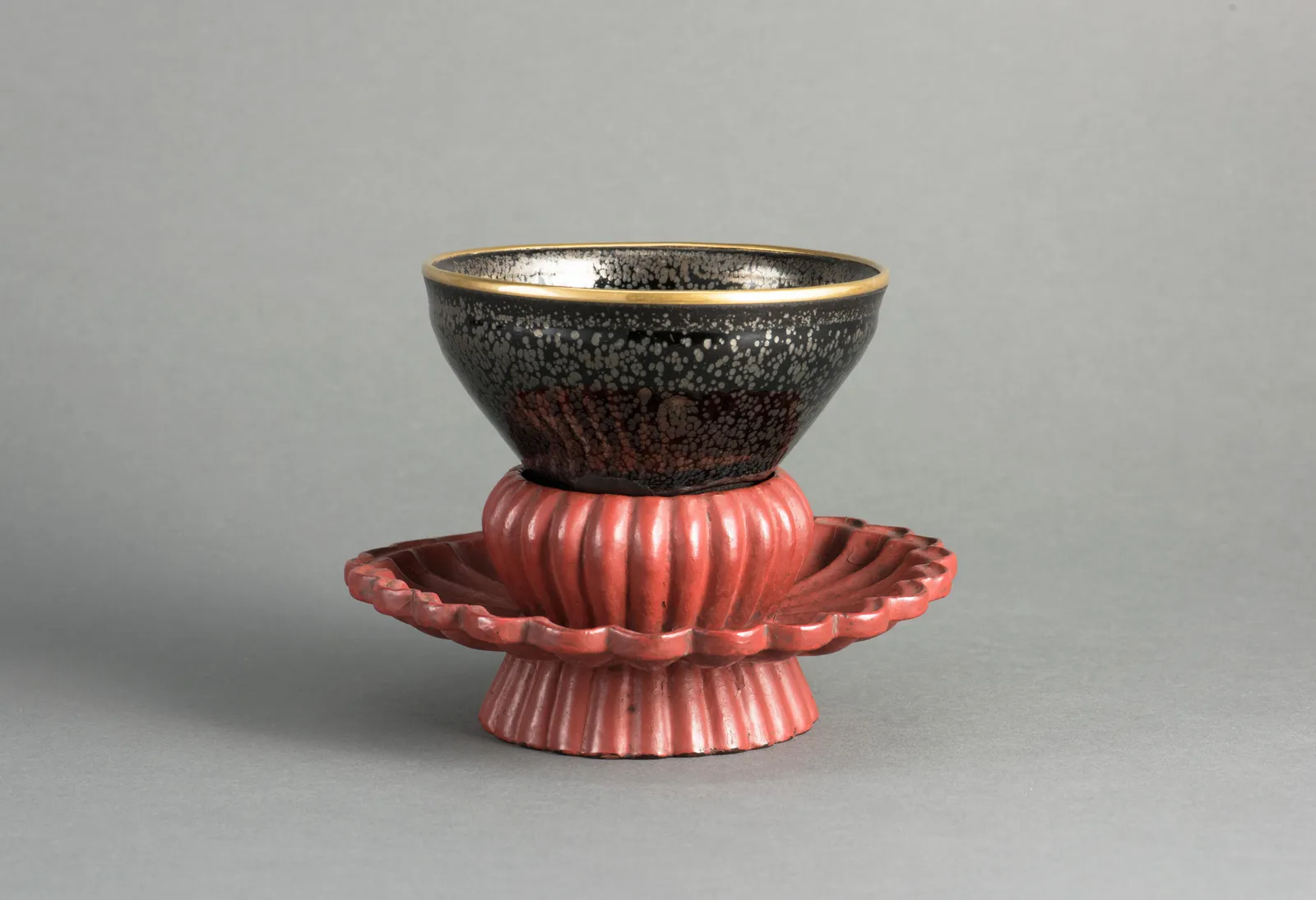 A Southern Song tea bowl with a black glaze scattered with silvery oil-spot markings and a gold rim, resting on a fluted red lacquer stand against a plain grey background