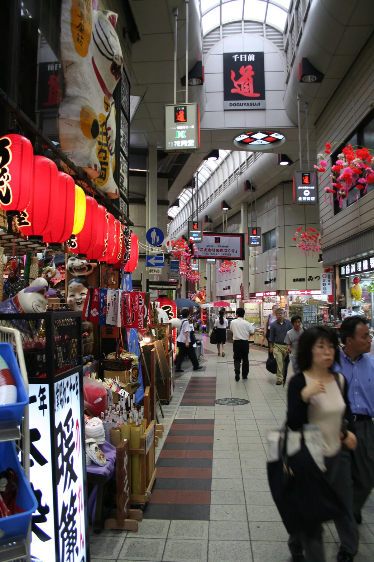 The Doguyasuji arcade in Osaka under its roof, a narrow covered lane of kitchenware shops with red paper lanterns and beckoning-cat figures stacked at the shopfronts and a lit sign overhead